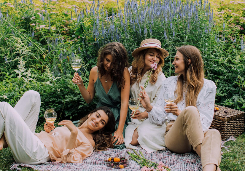 Quatre jeunes femmes souriantes profitant d'un pique-nique chic dans un jardin verdoyant avec des verres de vin blanc.